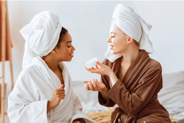 Dwie młode kobiety w szlafrokach i ręcznikach na głowie są zaciekawione kremem do twarzy, który trzyma w dłoni jedna z nich